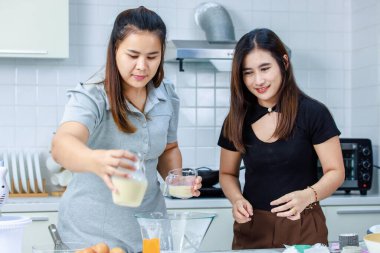 Asyalı profesyonel kadın pastane şefi ev hanımı ve arkadaşı sütçülerin taze sütü, dekore edilmiş mutfakta cam bardağa doldurup lezzetli bir tatlı pişirmeye yardım ediyorlar..
