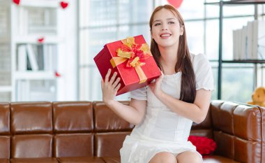 Beyaz elbiseli Asyalı güzel bayan manken kalp şeklinde koltukta oturmuş gülümsüyor. Oturma odasında sevgililer gününü kutluyor..