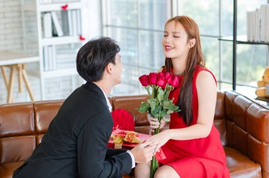 Resmi siyah takım elbiseli Asyalı yakışıklı erkek arkadaş kırmızı elbiseli, romantik dekore edilmiş oturma odasında oturan güzel kız arkadaşa elmas nişan yüzüğü takıyor..