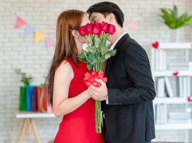 Resmi siyah takım elbiseli Asyalı genç erkek arkadaş ve kırmızı elbiseli güzel bayan kız arkadaş ellerinde kırmızı güller, çiçek buketiyle romantik bir salonda öpüşüyorlar..