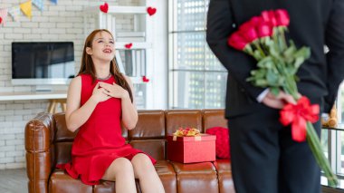Asyalı, genç, güzel, şaşkın, kırmızı elbiseli kız arkadaş rahat koltukta gülümserken tanınmayan erkek arkadaş, sevgililer gününde romantik dekore edilmiş oturma odasının arkasında çiçek buketi tutuyor..