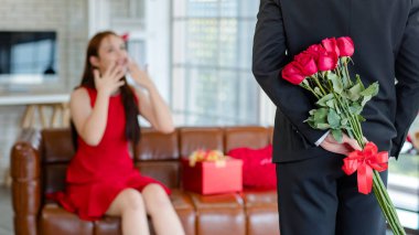 Asyalı, genç, güzel, şaşkın, kırmızı elbiseli kız arkadaş rahat koltukta gülümserken tanınmayan erkek arkadaş, sevgililer gününde romantik dekore edilmiş oturma odasının arkasında çiçek buketi tutuyor..