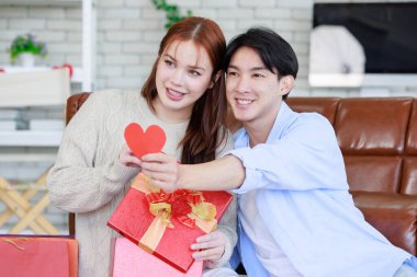 Asyalı genç ve güzel bayan kız arkadaş oturmuş gülümsüyor. Kutusuz kırmızı hediye paketini açıyor. Sevgililer gününde romantik dekore edilmiş oturma odasındaki yakışıklı erkek arkadaştan samimi bir kanepeye sarılmış..