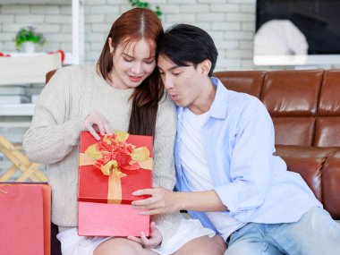 Asyalı genç ve güzel bayan kız arkadaş oturmuş gülümsüyor. Kutusuz kırmızı hediye paketini açıyor. Sevgililer gününde romantik dekore edilmiş oturma odasındaki yakışıklı erkek arkadaştan samimi bir kanepeye sarılmış..