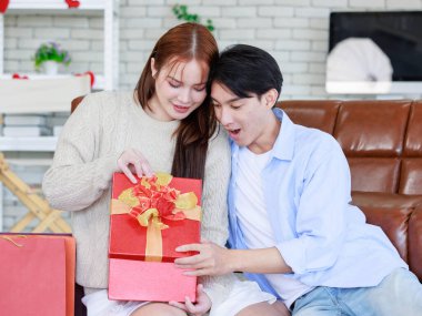 Asyalı genç ve güzel bayan kız arkadaş oturmuş gülümsüyor. Kutusuz kırmızı hediye paketini açıyor. Sevgililer gününde romantik dekore edilmiş oturma odasındaki yakışıklı erkek arkadaştan samimi bir kanepeye sarılmış..