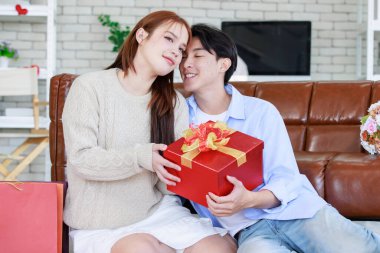 Asyalı genç ve güzel bayan kız arkadaş oturmuş gülümsüyor. Kutusuz kırmızı hediye paketini açıyor. Sevgililer gününde romantik dekore edilmiş oturma odasındaki yakışıklı erkek arkadaştan samimi bir kanepeye sarılmış..