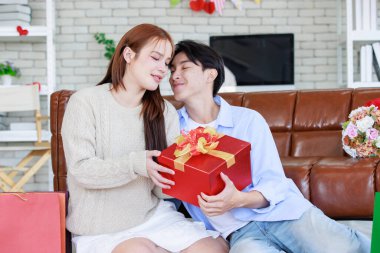 Asyalı genç ve güzel bayan kız arkadaş oturmuş gülümsüyor. Kutusuz kırmızı hediye paketini açıyor. Sevgililer gününde romantik dekore edilmiş oturma odasındaki yakışıklı erkek arkadaştan samimi bir kanepeye sarılmış..