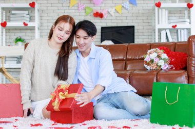Asyalı genç ve güzel bayan kız arkadaş oturmuş gülümsüyor. Kutusuz kırmızı hediye paketini açıyor. Sevgililer gününde romantik dekore edilmiş oturma odasındaki yakışıklı erkek arkadaştan samimi bir kanepeye sarılmış..