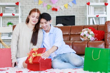 Asyalı genç ve güzel bayan kız arkadaş oturmuş gülümsüyor. Kutusuz kırmızı hediye paketini açıyor. Sevgililer gününde romantik dekore edilmiş oturma odasındaki yakışıklı erkek arkadaştan samimi bir kanepeye sarılmış..
