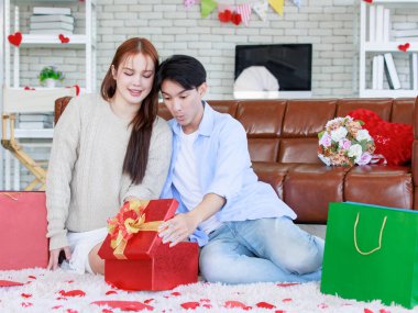 Asyalı genç ve güzel bayan kız arkadaş oturmuş gülümsüyor. Kutusuz kırmızı hediye paketini açıyor. Sevgililer gününde romantik dekore edilmiş oturma odasındaki yakışıklı erkek arkadaştan samimi bir kanepeye sarılmış..