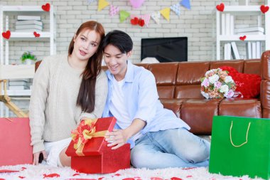 Asyalı genç ve güzel bayan kız arkadaş oturmuş gülümsüyor. Kutusuz kırmızı hediye paketini açıyor. Sevgililer gününde romantik dekore edilmiş oturma odasındaki yakışıklı erkek arkadaştan samimi bir kanepeye sarılmış..