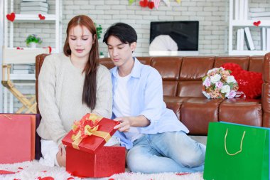 Asyalı genç ve güzel bayan kız arkadaş oturmuş gülümsüyor. Kutusuz kırmızı hediye paketini açıyor. Sevgililer gününde romantik dekore edilmiş oturma odasındaki yakışıklı erkek arkadaştan samimi bir kanepeye sarılmış..