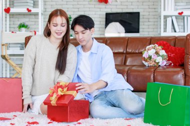 Asyalı genç ve güzel bayan kız arkadaş oturmuş gülümsüyor. Kutusuz kırmızı hediye paketini açıyor. Sevgililer gününde romantik dekore edilmiş oturma odasındaki yakışıklı erkek arkadaştan samimi bir kanepeye sarılmış..