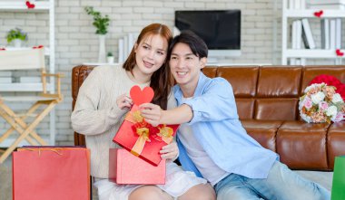 Asyalı genç ve güzel bayan kız arkadaş oturmuş gülümsüyor. Kutusuz kırmızı hediye paketini açıyor. Sevgililer gününde romantik dekore edilmiş oturma odasındaki yakışıklı erkek arkadaştan samimi bir kanepeye sarılmış..