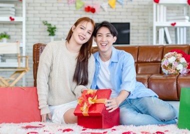 Asyalı genç ve güzel bayan kız arkadaş oturmuş gülümsüyor. Kutusuz kırmızı hediye paketini açıyor. Sevgililer gününde romantik dekore edilmiş oturma odasındaki yakışıklı erkek arkadaştan samimi bir kanepeye sarılmış..
