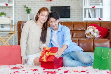 Asyalı genç ve güzel bayan kız arkadaş oturmuş gülümsüyor. Kutusuz kırmızı hediye paketini açıyor. Sevgililer gününde romantik dekore edilmiş oturma odasındaki yakışıklı erkek arkadaştan samimi bir kanepeye sarılmış..