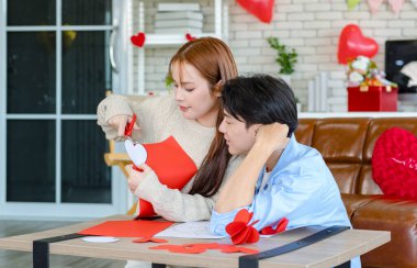 Asyalı genç, yakışıklı erkek arkadaş kanepede oturmuş gülümsüyor. Elinde kırmızı kalp şeklinde kağıt olan güzel kız arkadaşına sarılıyor. Sevgililer Günü 'nü kutlamak için romantik dekore edilmiş oturma odasını kesiyor..