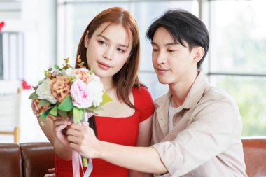 Asyalı genç, güzel bayan kız arkadaş ve elinde çiçek buketi olan yakışıklı erkek arkadaş sevgililer gününde evde oturma odasında sevgili olarak sırıtırken kameraya gülümsüyorlar..
