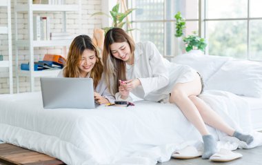 İki mutlu Asyalı güzel bayan kozmetik, cilt güzellik ürünlerine yüz bakımı yapar, dizüstü bilgisayar seyreden kadın, beyaz bir yatakta dinlenir, ergen ya da evdeki arkadaş yaşam tarzını kavrar..