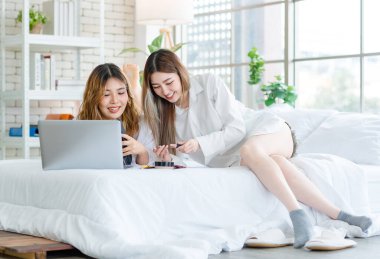 İki mutlu Asyalı güzel bayan kozmetik, cilt güzellik ürünlerine yüz bakımı yapar, dizüstü bilgisayar seyreden kadın, beyaz bir yatakta dinlenir, ergen ya da evdeki arkadaş yaşam tarzını kavrar..