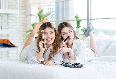 Beyaz bir yatak odasında uzanmış, makyaj yapan iki güzel Asyalı kadın gülümsemesi. Çekici bir kadın, güzellik ürünleri denerken eğleniyor, ergen ya da kız arkadaş yaşam tarzını benimsiyor..