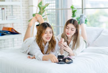 Two happy beautiful Asian female smile lying relaxed on a white bedroom, putting makeup. Attractive woman having fun together trying beauty products, Concept teenage or girlfriends lifestyle at home. clipart