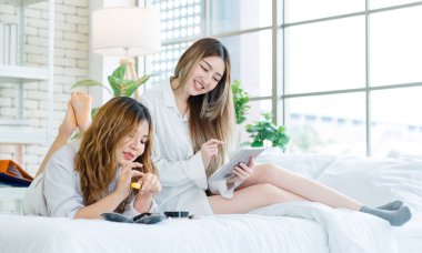 İki mutlu Asyalı güzel bayan kozmetik cilt bakım ürünleri, beyaz bir yatakta rahatlamış tablet izleyen güzel bir kadın, genç ya da evdeki arkadaş yaşam tarzı kavramını kavrıyor..