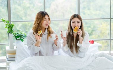 Two beautiful people happy Asian females drinking hot coffee or tea with bread or cake. Pretty woman laughing and talking relaxed funny on a white bed, Concept teenage or friends lifestyle at home. clipart