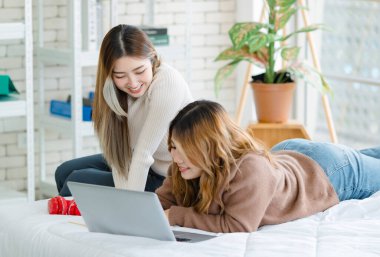 İki mutlu Asyalı güzel kız arkadaş mutlu, gülümseyen bir bilgisayar izliyorlar, beyaz bir yatakta rahatça uzanıyorlar, eğleniyorlar, gençlerin ya da arkadaşlarının evdeki yaşam tarzlarını kavrıyorlar.