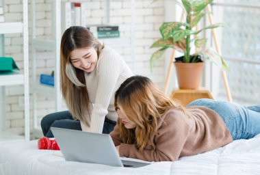 İki mutlu Asyalı güzel kız arkadaş mutlu, gülümseyen bir bilgisayar izliyorlar, beyaz bir yatakta rahatça uzanıyorlar, eğleniyorlar, gençlerin ya da arkadaşlarının evdeki yaşam tarzlarını kavrıyorlar.