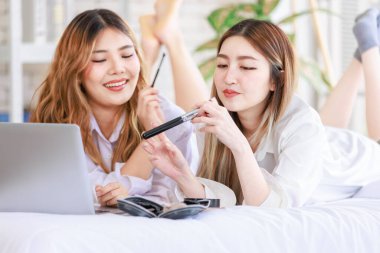 İki mutlu Asyalı güzel bayan kozmetik, cilt güzellik ürünlerine yüz bakımı yapar, dizüstü bilgisayar seyreden kadın, beyaz bir yatakta dinlenir, ergen ya da evdeki arkadaş yaşam tarzını kavrar..