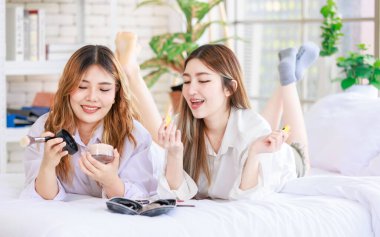 Beyaz bir yatak odasında uzanmış, makyaj yapan iki güzel Asyalı kadın gülümsemesi. Çekici bir kadın, güzellik ürünleri denerken eğleniyor, ergen ya da kız arkadaş yaşam tarzını benimsiyor..