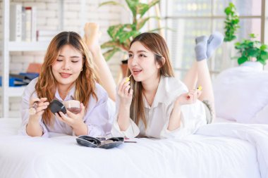 Beyaz bir yatak odasında uzanmış, makyaj yapan iki güzel Asyalı kadın gülümsemesi. Çekici bir kadın, güzellik ürünleri denerken eğleniyor, ergen ya da kız arkadaş yaşam tarzını benimsiyor..