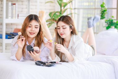 Beyaz bir yatak odasında uzanmış, makyaj yapan iki güzel Asyalı kadın gülümsemesi. Çekici bir kadın, güzellik ürünleri denerken eğleniyor, ergen ya da kız arkadaş yaşam tarzını benimsiyor..