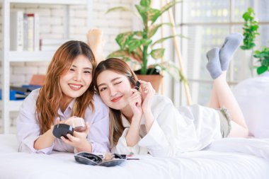 Beyaz bir yatak odasında uzanmış, makyaj yapan iki güzel Asyalı kadın gülümsemesi. Çekici bir kadın, güzellik ürünleri denerken eğleniyor, ergen ya da kız arkadaş yaşam tarzını benimsiyor..