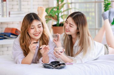Beyaz bir yatak odasında uzanmış, makyaj yapan iki güzel Asyalı kadın gülümsemesi. Çekici bir kadın, güzellik ürünleri denerken eğleniyor, ergen ya da kız arkadaş yaşam tarzını benimsiyor..