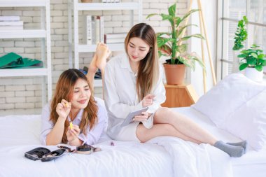 İki mutlu Asyalı güzel bayan kozmetik cilt bakım ürünleri, beyaz bir yatakta rahatlamış tablet izleyen güzel bir kadın, genç ya da evdeki arkadaş yaşam tarzı kavramını kavrıyor..
