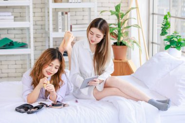 İki mutlu Asyalı güzel bayan kozmetik cilt bakım ürünleri, beyaz bir yatakta rahatlamış tablet izleyen güzel bir kadın, genç ya da evdeki arkadaş yaşam tarzı kavramını kavrıyor..