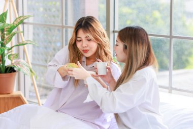 Two beautiful people happy Asian females drinking hot coffee or tea with bread or cake. Pretty woman laughing and talking relaxed funny on a white bed, Concept teenage or friends lifestyle at home. clipart