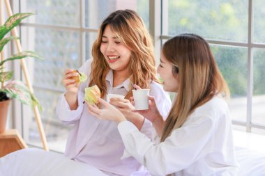 Two beautiful people happy Asian females drinking hot coffee or tea with bread or cake. Pretty woman laughing and talking relaxed funny on a white bed, Concept teenage or friends lifestyle at home. clipart