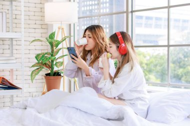 Two beautiful people happy Asian females drinking hot coffee or tea with bread or cake. Pretty woman laughing and talking relaxed funny on a white bed, Concept teenage or friends lifestyle at home. clipart