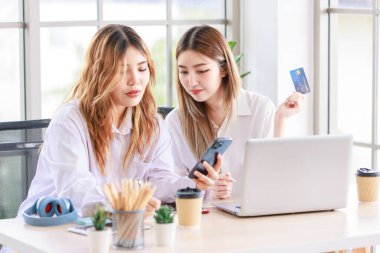 Two happy women, a beautiful Asian female using a computer laptop smiling while shopping online, and a credit card in the living room. Concept teenage or friends, time together lifestyle at home. clipart