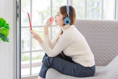 Asyalı genç kadın, güzel bayan, rahatla. Kulaklıkla müzik dinle. Oturma odasının önündeki modern koltukta bilgisayar tableti kullanarak otur. Gençlerin Yaşam Tarzı Hobisi Konsepti
