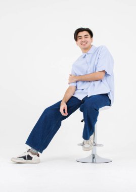 Asyalı erkek özgüveninin portresi yakışıklı yakışıklı, mavi yakalı gömlekli ve kot pantolonlu beyaz arka plan stüdyosundaki sandalyede oturan rahat görünüşlü, izole bir genç.