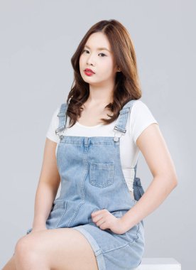 Asyalı sevimli, neşeli, kot pantolon giyen genç bir kızın portresi mutlu ve tatlı gülüşlü şık bir model kadın beyaz arka planda, gençlik ve gençlik modasında izole bir şekilde poz veren kutunun üzerinde oturuyor..