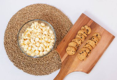 Tahta kesme tahtasına yerleştirilmiş çikolatalı kurabiyeler ve beyaz arka planda cam klozette patlamış mısır, parti yemeği konsepti..