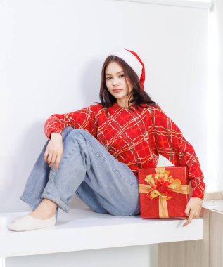 Happy Asian beautiful woman wearing santa hat red long sleeve knitted sweater sitting holding present gift box funny smiling behind white background, Christmas Eve Happy New Year's Party celebration. clipart