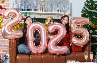 İki mutlu, genç, güzel Asyalı kadın, 2025 numaralı gül rengi balonları tutan sevimli arkadaşlar koltukta oturmuş Noel ağacı süslemenin tadını çıkarıyorlar. Yeni yıl partisi kutlaması..