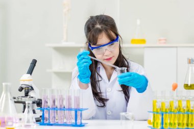 Asyalı genç kadın laboratuvar asistanı profesyonel bilim adamı beyaz laboratuvar önlüğü ve koruyucu gözlüklerle kimya laboratuarında deney tüpleri üzerinde araştırma yapıyor.
