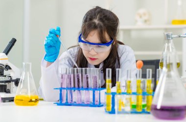 Asyalı genç kadın laboratuvar asistanı profesyonel bilim adamı beyaz laboratuvar önlüğü ve koruyucu gözlüklerle kimya laboratuarında deney tüpleri üzerinde araştırma yapıyor.